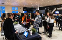 Ihmisiä rekisteröitymässä konferenssiin modernissa sisätilassa. Pöydällä on nimikortteja ja kannettava tietokone. Osallistujat näyttävät iloisilta ja keskustelevat keskenään. Taustalla näkyy pukukaappeja ja iso näyttö.