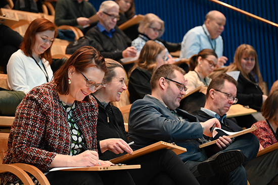 Hymyileviä seminaarin osallistujia luentosalissa kirjoittamassa muistiinpanoja.